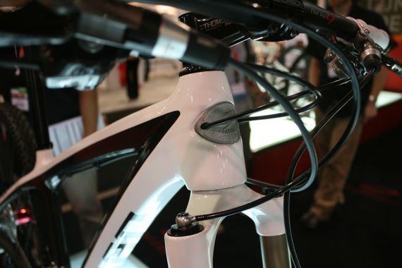 Close-up view of a white mountain bike frame, highlighting the head tube and cable routing. The image features a sleek design with a distinct logo on the head tube and components such as the handlebars and fork in the background, set against a blurred indoor display environment.