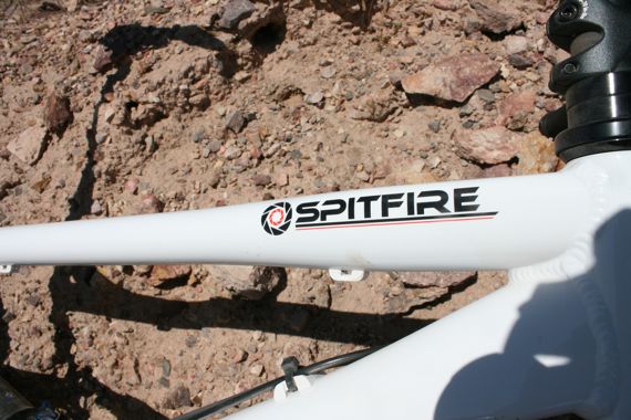 Close-up view of a white bicycle frame featuring the "Spitfire" logo on the top tube, set against a rocky terrain.