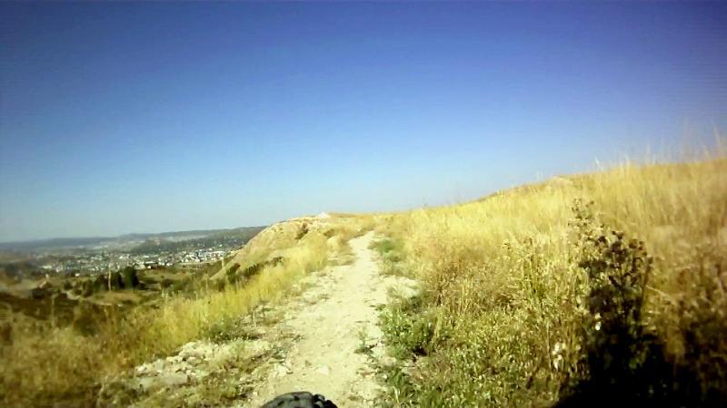 A dirt pathway winding through dry grass and gentle hills under a clear blue sky, with a distant view of a city beyond the landscape. Quarry Mesa mountain bike trail.