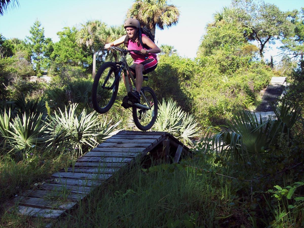 A young girl wearing a helmet and athletic clothing is airborne while riding a mountain bike over a wooden ramp in a wooded area. Lush greenery and palm-like plants surround the scene, highlighting a vibrant outdoor environment. Jonathan Dickinson State Park mountain bike trail.