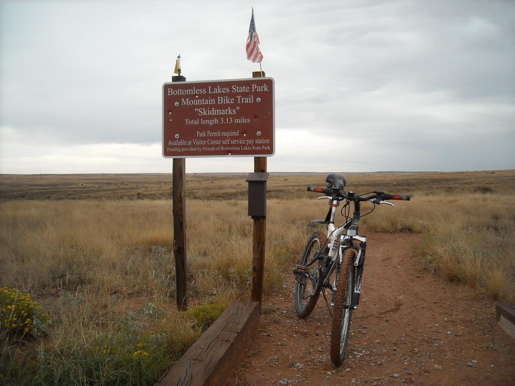 A mountain bike resting next to a sign for the Bottomless Lakes State Park Mountain Bike Trail named "Skidmarks." The sign indicates the trail's total length of 3.13 miles and notes that a park permit is required. The background features a wide, open landscape with tall grass and a cloudy sky. Skidmarks mountain bike trail.
