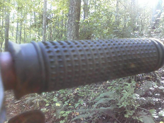 Close-up view of a textured black bicycle handlebar grip set against a blurred background of green foliage and trees, suggesting an outdoor setting. The image captures the grip's rubber material and pattern, emphasizing its design for comfort and control. Heritage Park mountain bike trail.