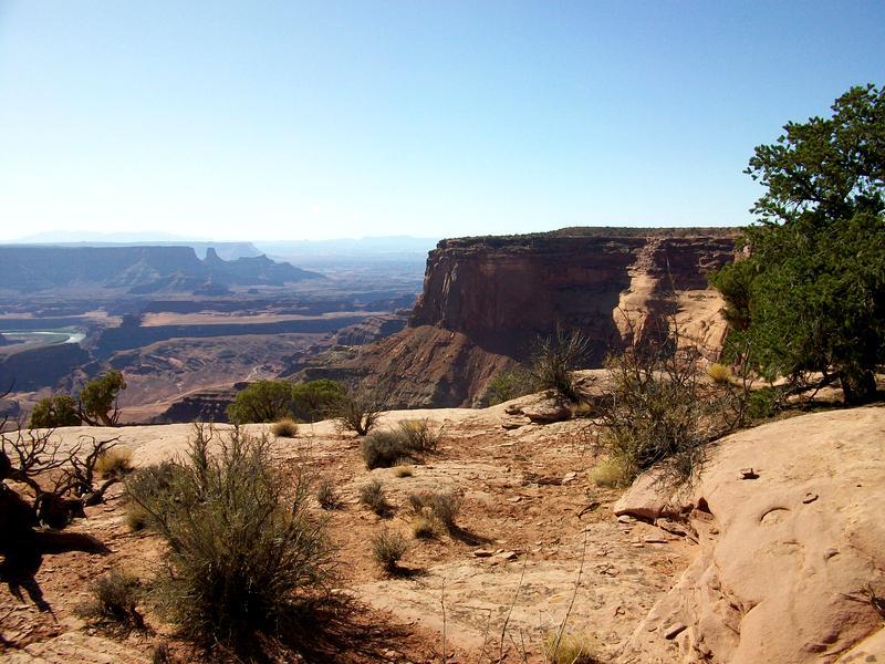Alt text: A panoramic view of a rugged canyon landscape with rocky cliffs and sparse vegetation, under a clear blue sky. The scene captures the contrasting colors of the earth and the distant mountains, showcasing a natural desert environment. Dead Horse Point State Park mountain bike trail.
