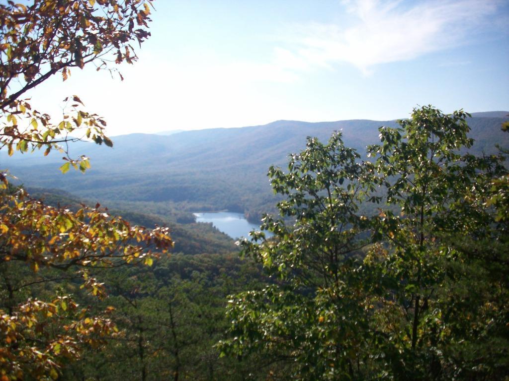 A scenic view of rolling mountains and a serene lake surrounded by lush greenery, with hints of autumn foliage in the trees under a clear blue sky. Douthat State Park mountain bike trail.