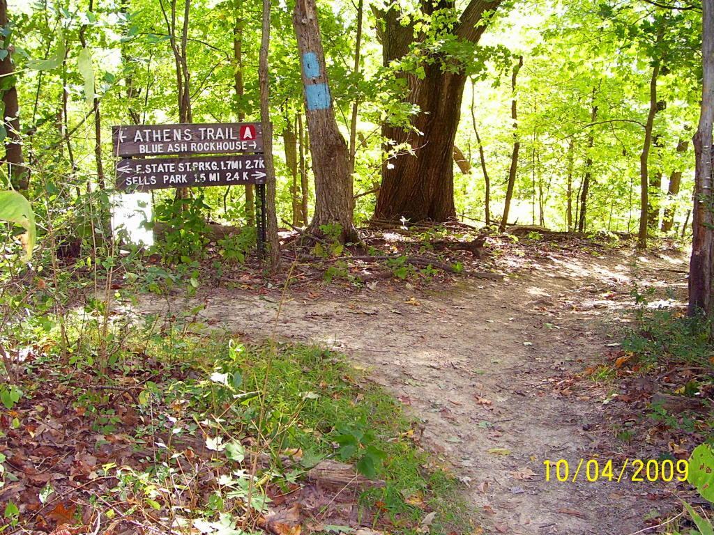 A dirt path leading into a wooded area, with a signpost displaying directions for the Athens Trail, Blue Ash Rockhouse, and nearby locations, under dappled sunlight filtering through the trees. The sign indicates distances to East State Street Park and Sells Park. Strouds Run State Park mountain bike trail.