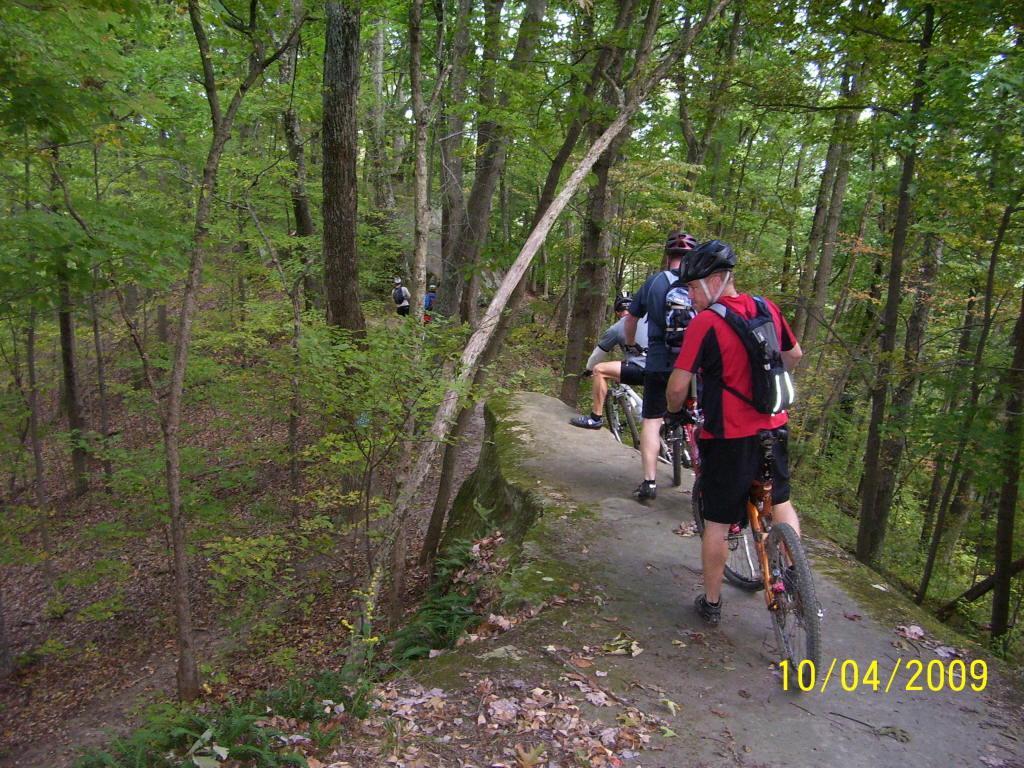 A group of mountain bikers riding along a narrow trail in a wooded area, surrounded by lush green trees and foliage. The path is rocky and uneven, indicating a challenging terrain. The cyclists are wearing helmets and athletic gear, showing their engagement in outdoor activities. Strouds Run State Park mountain bike trail.