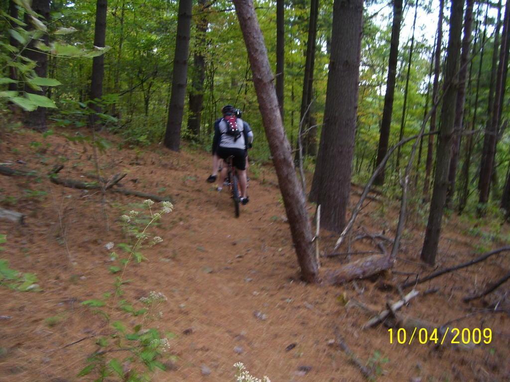 A cyclist riding a mountain bike through a forested trail surrounded by tall trees and pine needles on the ground. The scene depicts a blend of greenery and sunlight filtering through the foliage. The date of the photo, October 4, 2009, is visible in the bottom right corner. Strouds Run State Park mountain bike trail.