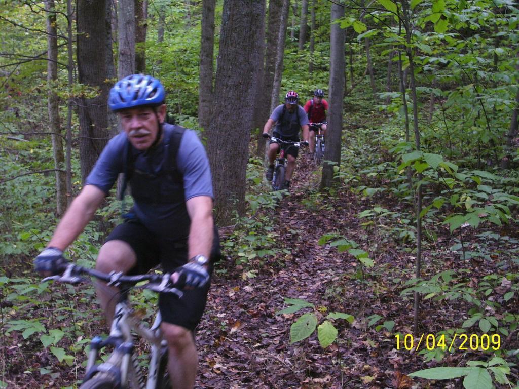 Three mountain bikers navigate a narrow, wooded trail covered in fallen leaves. The first rider, in the foreground, wears a blue helmet and dark cycling attire. The other two riders trail behind, appearing focused as they ride through the lush, green forest. Strouds Run State Park mountain bike trail.