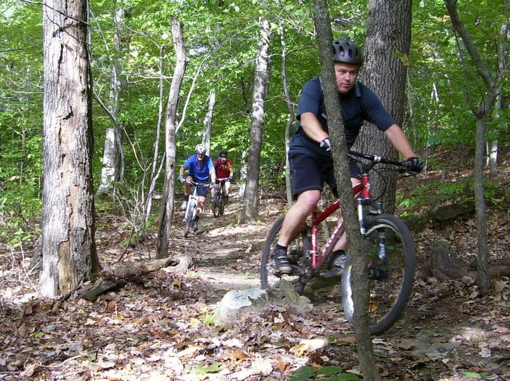 Three cyclists on a narrow, wooded trail surrounded by trees and fallen leaves, with sunlight filtering through the foliage. One cyclist is in the foreground, riding a red mountain bike, while the others are further back, wearing blue and red shirts. Strouds Run State Park mountain bike trail.
