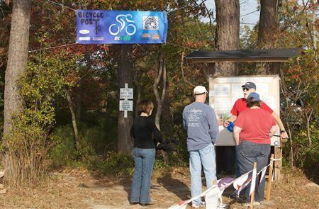 A group of three people gathered at a bicycle post information area, with a sign indicating bike trails above them. Two individuals stand in front of the informational board while a third person watches. The setting features trees and autumn foliage in the background. Bicycle Post Trails mountain bike trail.