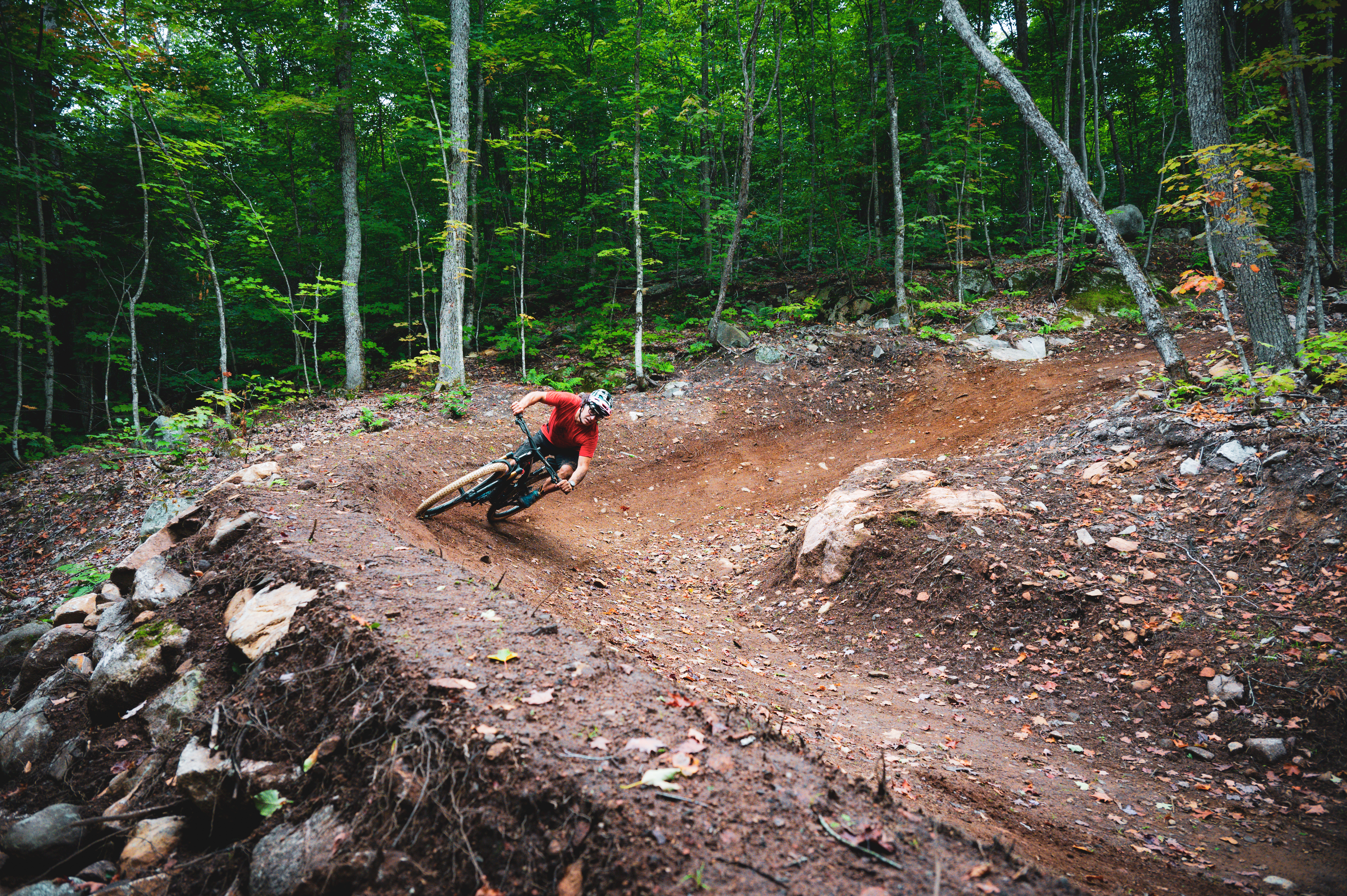 A cyclist in a red shirt leans into a curve on a dirt mountain biking trail surrounded by trees and rocky terrain. Hiawatha mountain bike trail.