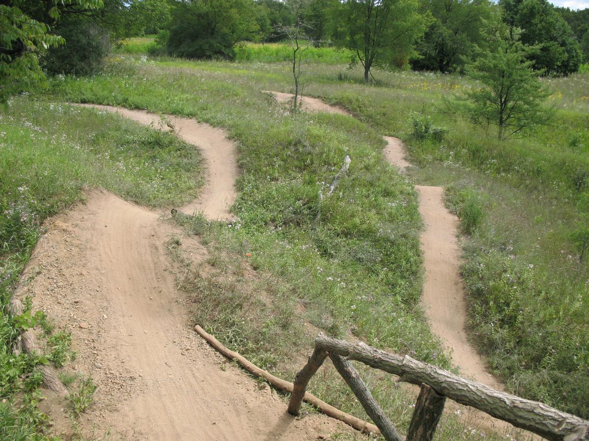A winding dirt bike trail through a grassy landscape, featuring several curves and gentle hills, surrounded by trees and wildflowers. Fort Custer Recreation Area mountain bike trail.