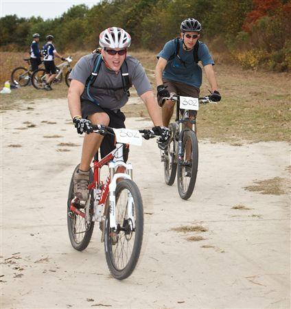 Two mountain bikers are racing on a dirt trail. One rider, wearing a helmet and sunglasses, is in the foreground, pushing ahead with a focused expression. The second rider trails behind, also equipped with a helmet and a race number on their shirt. In the background, several other cyclists are visible, and the landscape features autumn foliage. Bicycle Post Trails mountain bike trail.