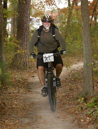 A mountain biker wearing a helmet and backpack rides along a dirt trail surrounded by trees, with fallen leaves scattered on the ground. The cyclist has a race number attached to his shorts and is focused on navigating the path. Bicycle Post Trails mountain bike trail.