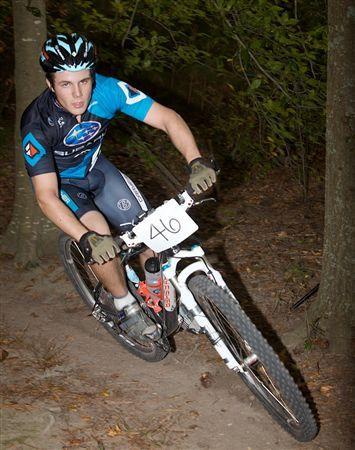 A young male mountain biker wearing a blue and black cycling jersey and helmet leans into a turn on a dirt trail surrounded by trees. He is focused and determined, riding a white mountain bike with a race number 46 displayed on the front. The setting suggests a competitive outdoor event. Bicycle Post Trails mountain bike trail.