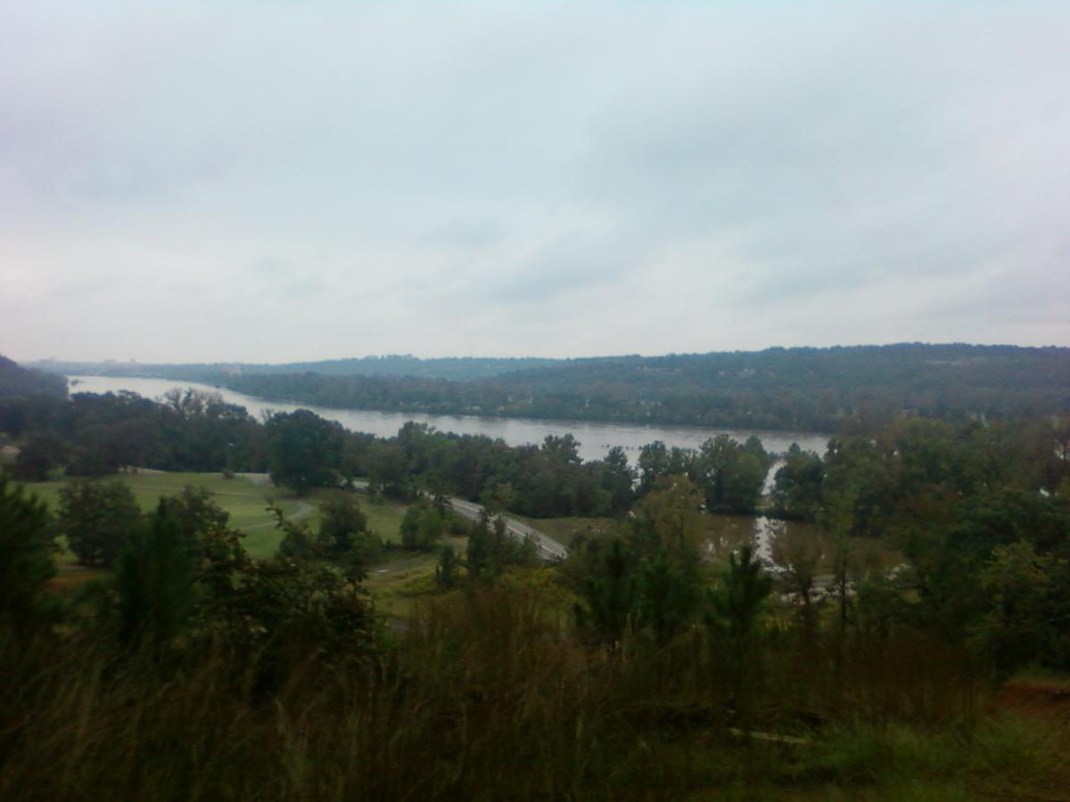 A misty view of a river winding through a lush landscape, with trees lining the banks and rolling hills in the background under a cloudy sky. Burns Park mountain bike trail.