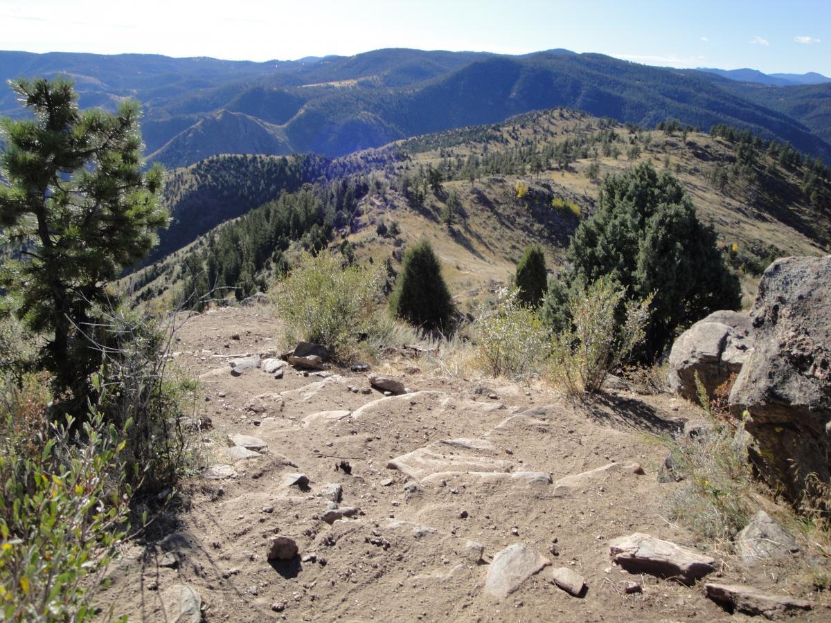 A scenic view of rolling mountains and valleys under a clear blue sky. The foreground features rocky terrain with patches of soil and sparse vegetation, including shrubs and a pine tree. In the background, layers of hills and wooded areas create a beautiful, natural landscape. Centennial Cone Park mountain bike trail.