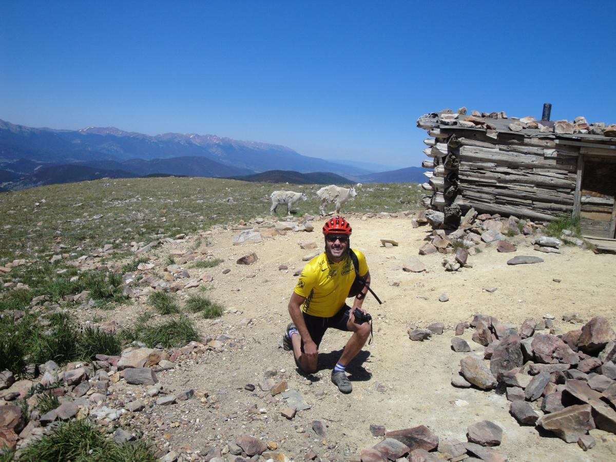 A cyclist in a bright yellow jersey and helmet kneels on rocky terrain near a rustic wooden cabin, set against a backdrop of mountains under a clear blue sky. Colorado Trail mountain bike trail.