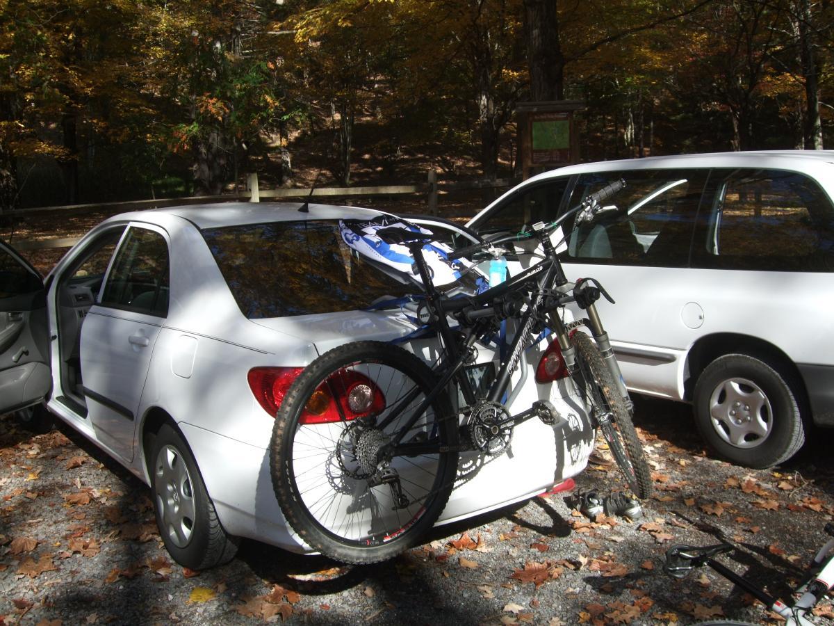 A white car parked among autumn foliage, with a mountain bike secured on its rear. The car's door is open, revealing the interior. Nearby, another vehicle is visible in the background, and the ground is covered with colorful autumn leaves. Douthat State Park mountain bike trail.