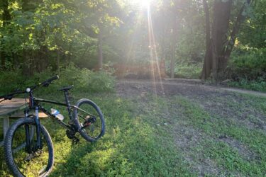 A mountain bike resting on a grassy area next to a wooden picnic table, surrounded by dense green foliage. The sunlight filters through the trees, creating a warm, inviting atmosphere. In the background, a path leads further into the woods. Creve Couer Park mountain bike trail.
