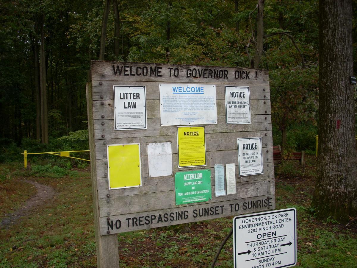 A wooden sign at the entrance of Governor Dick Park displaying various notices, including rules about littering, trespassing, and park hours. The sign states "Welcome to Governor Dick" at the top, with additional signs detailing park regulations and information about the environmental center's hours of operation. The background features wooded area and a path leading into the park. Trails At Mt. Gretna mountain bike trail.