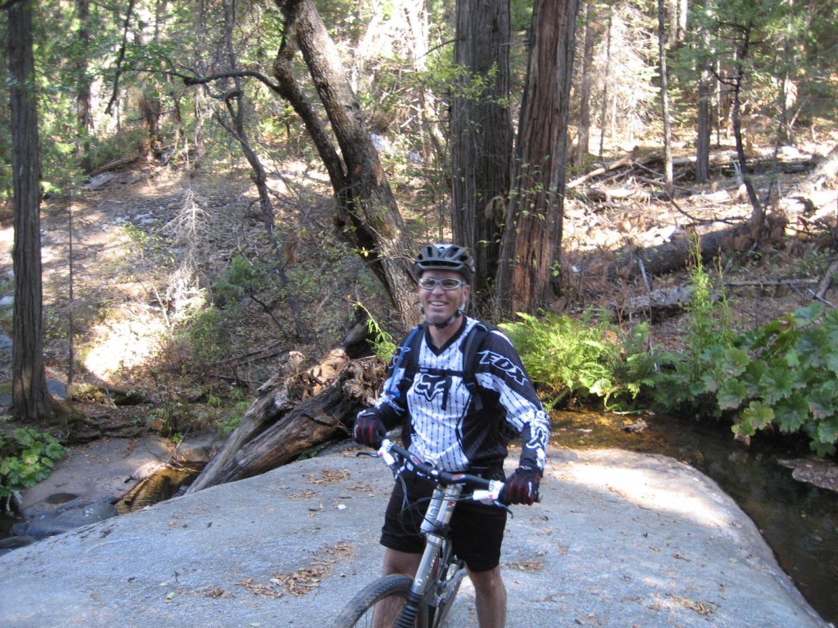 A person wearing a helmet and cycling gear stands on a rocky area next to a creek, surrounded by tall trees and lush greenery. They are smiling and holding a mountain bike, with sunlight filtering through the trees in the background. Top Of The World mountain bike trail.