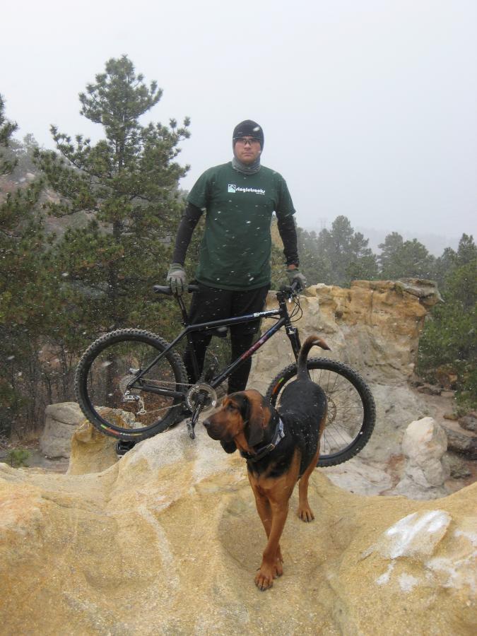 A person wearing a green shirt and black beanie stands on a rocky outcrop in a snowy landscape, next to a mountain bike. A brown and black dog stands nearby, looking curiously at the camera. Snow is gently falling, and pine trees can be seen in the background. Palmer Park mountain bike trail.