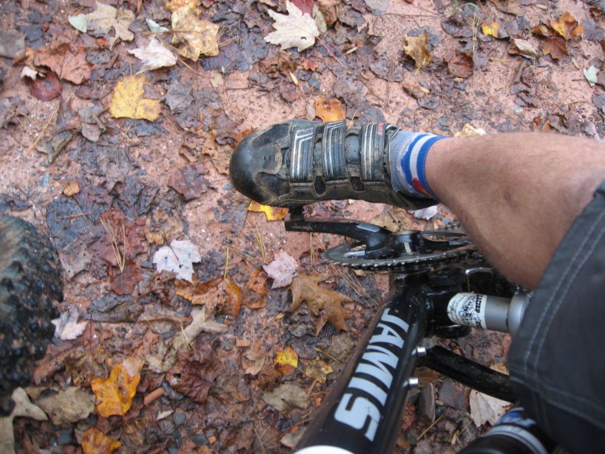 A cyclist's foot on a mountain bike pedal, surrounded by fallen leaves and dirt, with mud on the shoe and visible bike components. Bull / Jake Mountain mountain bike trail.