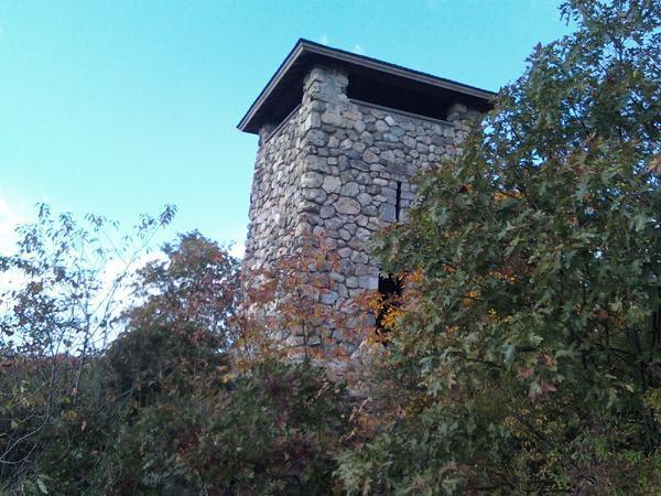 A stone tower partially obscured by trees and foliage against a blue sky. The tower features a flat roof and has large openings on one side, suggesting it may have been used for viewing or lookout purposes. Middlesex Fells Reservation mountain bike trail.