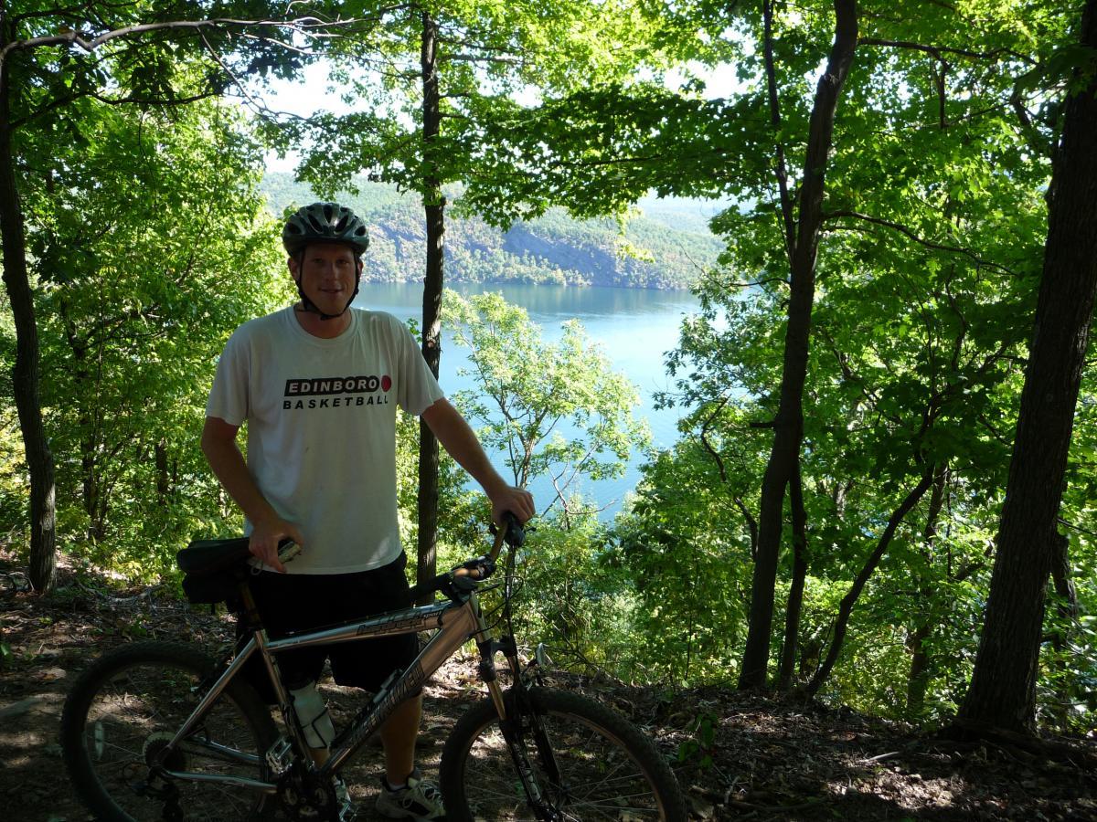 A person wearing a helmet and an Edinboro Basketball t-shirt stands next to a mountain bike, surrounded by lush green trees. In the background, a serene lake reflects the greenery, with rolling hills visible on the horizon under a clear blue sky. The image captures a moment of outdoor adventure in a natural setting. Allegrippis Trails mountain bike trail.