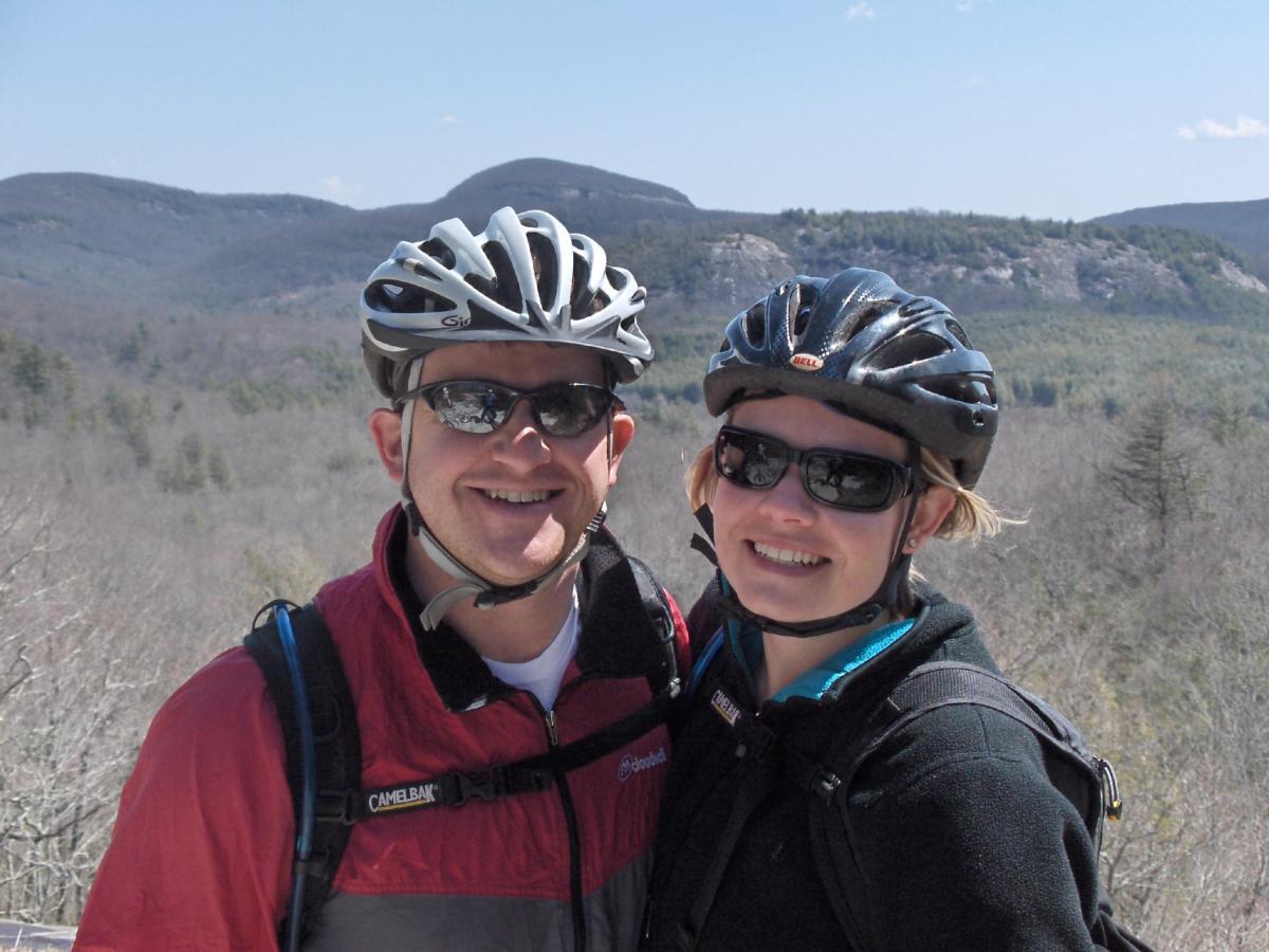 A smiling couple wearing cycling helmets and sunglasses, standing outdoors with a mountainous landscape in the background. They are dressed in outdoor gear, and the scene is bright with clear blue skies. Panthertown Valley mountain bike trail.