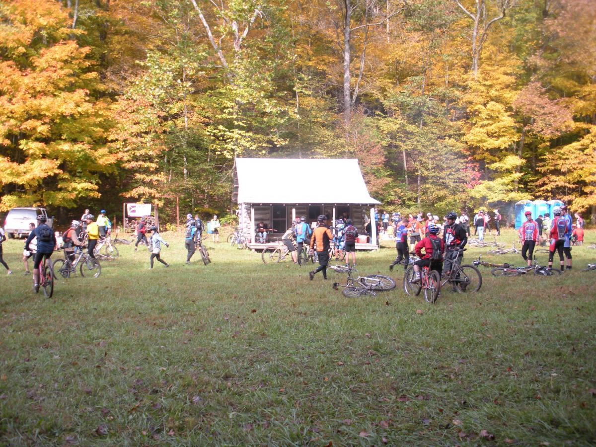 A group of cyclists gather in a grassy clearing surrounded by colorful autumn trees. They are near a log cabin, which serves as a hub for activity, while bicycles are parked around the area. Some participants are conversing and preparing for a ride, while others stand by the cabin. Portable restrooms are visible in the background. The scene captures the vibrant fall colors and the camaraderie of outdoor cycling events. Brown County Park mountain bike trail.