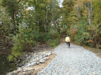 A person wearing a yellow jacket rides a bicycle along a gravel path beside a calm body of water, surrounded by trees with autumn foliage. Lake Maury mountain bike trail.