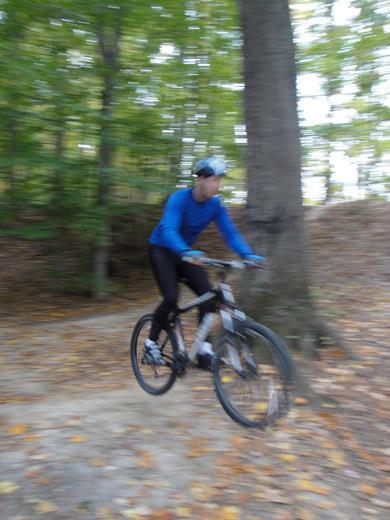A person in a blue long-sleeve shirt and black leggings riding a mountain bike on a wooded trail, captured in motion with a blurred background, indicating speed. Leaves cover the ground, and trees are visible in the surrounding area. Lake Maury mountain bike trail.