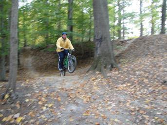 A person in a yellow jacket and helmet is riding a mountain bike and performing a jump on a dirt trail surrounded by trees with autumn leaves. Lake Maury mountain bike trail.