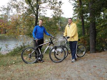 Two men standing next to their bicycles on a gravel path by a lake, surrounded by trees with autumn foliage. One man is wearing a blue long-sleeve shirt, and the other is in a yellow sweatshirt. The scene conveys a sense of outdoor activity and friendship. Lake Maury mountain bike trail.