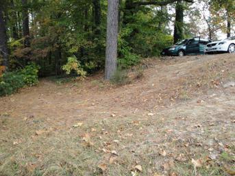 A grassy area with scattered autumn leaves, featuring a slight slope leading into a wooded area. In the background, two parked cars are visible among the trees. Lake Maury mountain bike trail.