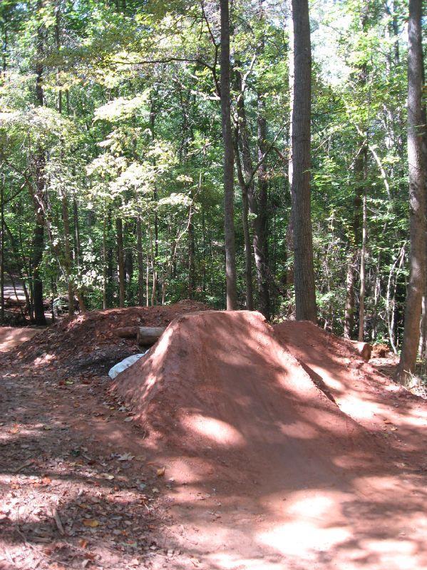 A dirt jump ramp located in a wooded area, surrounded by tall trees and dappled sunlight. The ramp is slightly elevated and has a smooth, curved surface, indicating use for BMX biking or similar sports. Leaf litter is visible on the ground, and a small log and white material can be seen near the base of the ramp. Big Creek mountain bike trail.