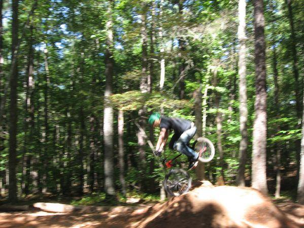 A cyclist performing a jump on a bike over a dirt ramp in a wooded area, with trees in the background. The image captures the motion with a blurred effect, emphasizing the dynamic movement of the rider. Big Creek mountain bike trail.