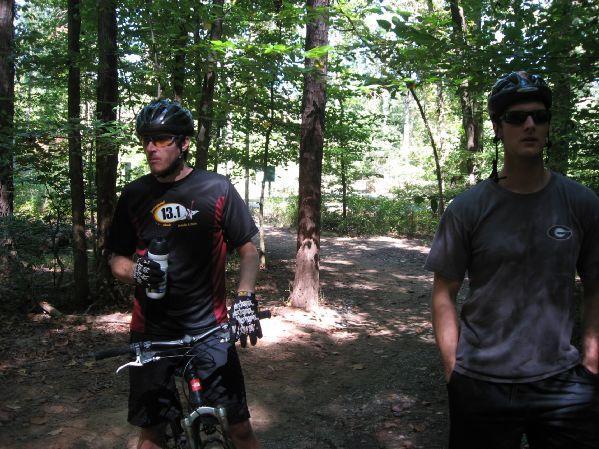 Two men in cycling gear are on a dirt path in a wooded area. One man, wearing a helmet and gloves, is holding a water bottle and standing next to a mountain bike. The other man, wearing sunglasses and a t-shirt, is standing nearby. Sunlight filters through the trees, creating a dappled effect on the ground. Big Creek mountain bike trail.