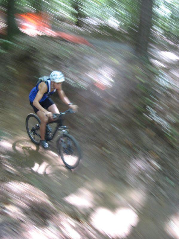 A cyclist in a blue tank top and helmet rides swiftly down a dirt path in a wooded area, with motion blur emphasizing speed and movement. Sunlight filters through the trees, creating dappled light on the ground. Big Creek mountain bike trail.
