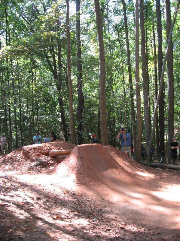 A dirt bike jump in a wooded area, with a group of spectators in the background observing the scene. Sunlight filters through the trees, creating a dappled light effect on the ground. Big Creek mountain bike trail.