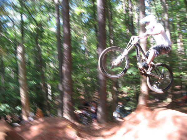 A mountain biker performing a jump off a dirt ramp in a wooded area, surrounded by trees, with motion blur conveying speed and excitement. Big Creek mountain bike trail.