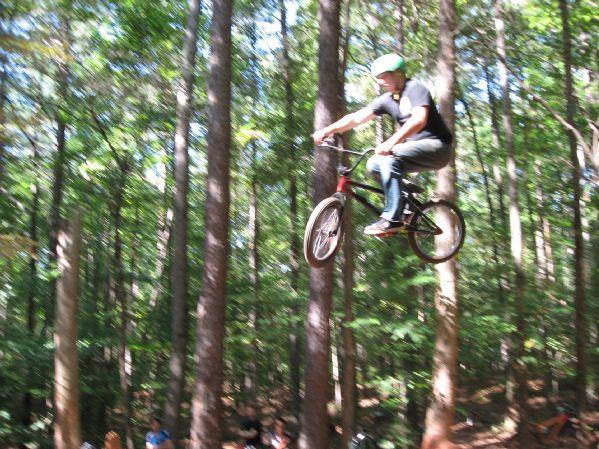 A person wearing a helmet jumps off a dirt ramp on a BMX bike, soaring through the air among tall trees in a forest. A small crowd can be seen in the background, watching the action. Big Creek mountain bike trail.
