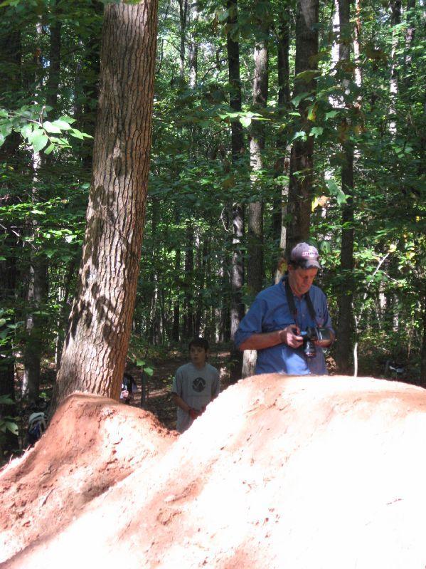 An older man holding a camera stands in a wooded area, focused on taking a photograph. In the background, a young boy watches. The scene features dirt mounds, likely made for biking or jumping, surrounded by tall trees with green foliage. Big Creek mountain bike trail.