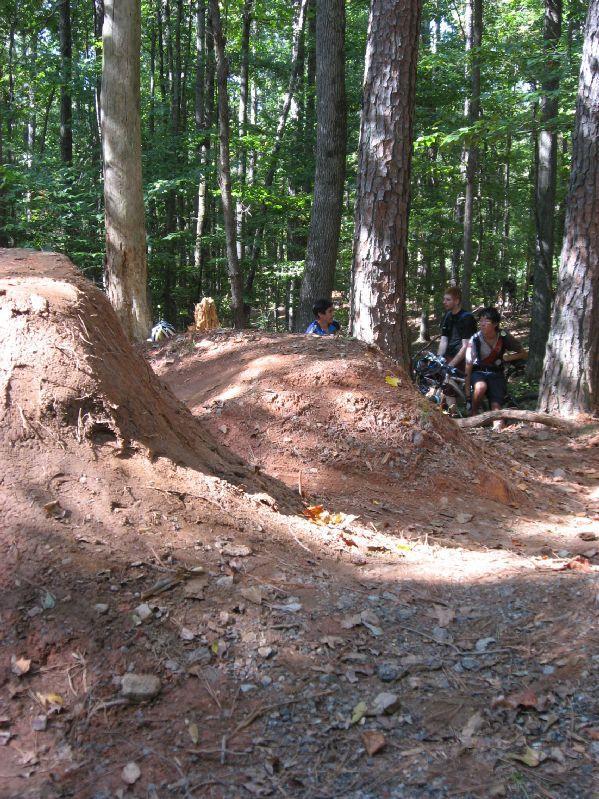 A dirt bike jump surrounded by trees in a wooded area, with two people sitting nearby on the ground, resting. Sunlight filters through the leaves, illuminating the scene. Big Creek mountain bike trail.
