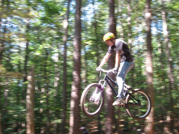 A mountain biker in mid-air performing a jump on a forest trail, wearing a yellow helmet and casual attire, surrounded by tall trees and dappled sunlight. Big Creek mountain bike trail.