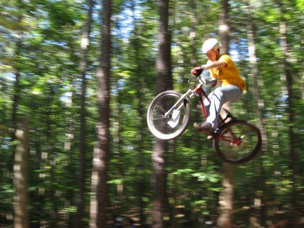 A cyclist in mid-air performing a jump on a mountain bike, surrounded by tall trees in a lush forest. The scene conveys speed and motion, with a blurred background emphasizing the dynamic action. The rider is wearing a helmet and a yellow shirt. Big Creek mountain bike trail.
