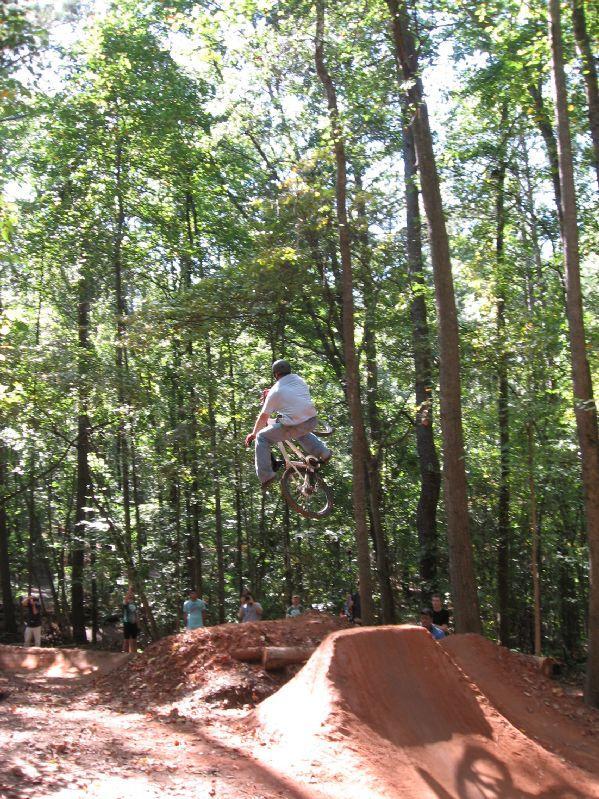 A cyclist performing a jump on a dirt bike at a forested BMX park, surrounded by spectators. The sun filters through the trees, creating a lively outdoor scene. Big Creek mountain bike trail.