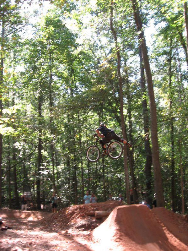 A cyclist mid-air performing a jump on a mountain bike in a wooded area. The scene captures the moment as the biker soars over a dirt ramp, surrounded by tall trees and a cheering crowd in the background. Big Creek mountain bike trail.